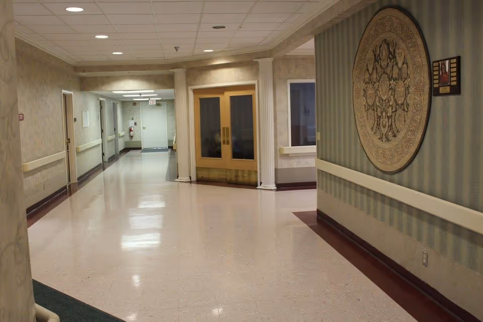 A clean, well-lit hallway in a healthcare facility with beige walls, a polished floor, and double doors at the end. The hallway features handrails on both sides, a decorative round wall hanging, and a small framed picture on the right wall.