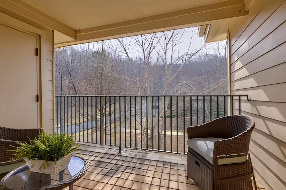 Covered balcony with wicker chairs, a small glass table holding a potted plant, and a metal railing overlooking trees.