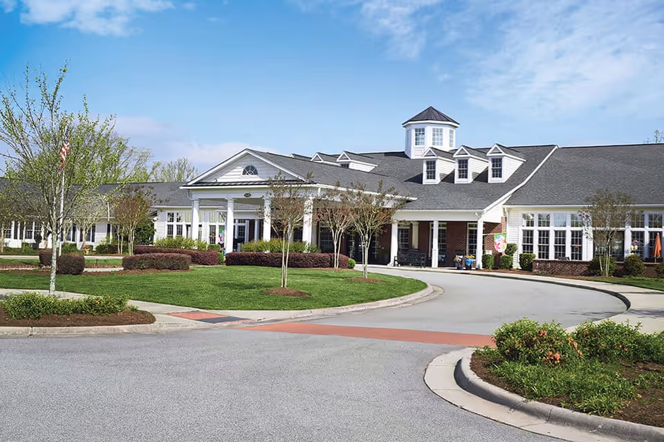 Exterior view of a single-story senior living facility building with a covered entrance, multiple windows, and a circular driveway surrounded by landscaped greenery and small trees under a partly cloudy sky.