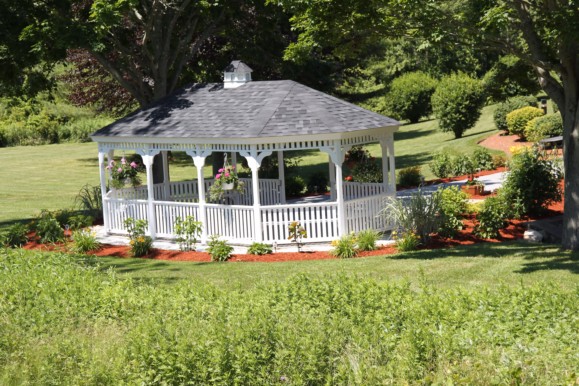 A white wooden gazebo with a gray shingled roof situated in a well-maintained garden area with green grass, bushes, and trees surrounding it. Hanging flower pots with pink flowers are attached to the gazebo, and there is a paved walkway nearby.