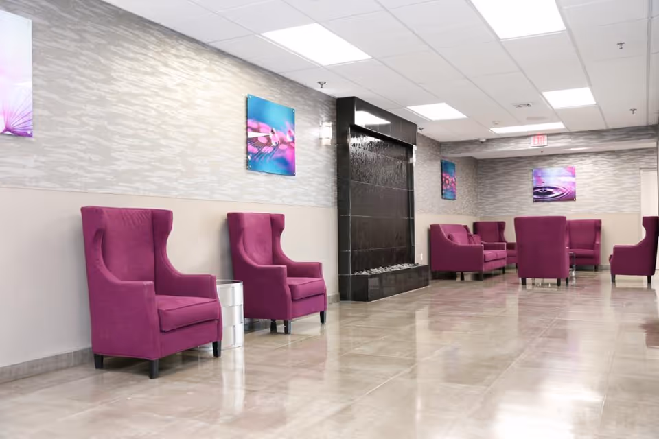 A spacious waiting or lounge area with multiple purple upholstered armchairs arranged along the walls and around a central space. The room features a modern black water feature on one wall, light gray patterned wallpaper, and several colorful abstract art pieces. The floor is tiled with a glossy finish, and the ceiling has recessed lighting panels.