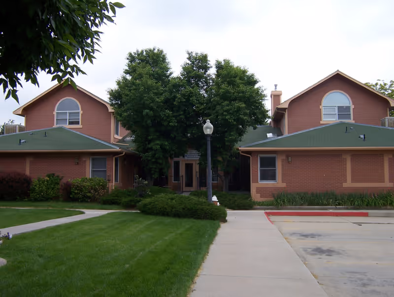 Front view of a two-story brick senior living building with green roofs, a central lamp post and walkway, and trees and lawn in the foreground.