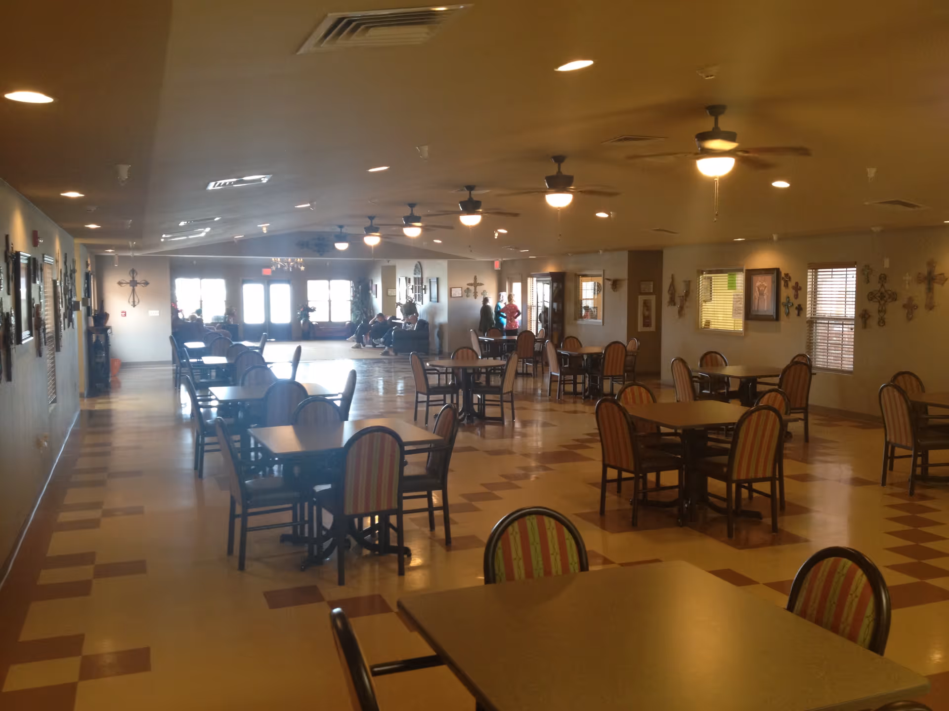 A spacious dining room with multiple tables and chairs arranged neatly. The room has ceiling fans with lights, windows with blinds, and walls decorated with various crosses and framed pictures. There are a few people visible in the background near the entrance and sitting on benches.