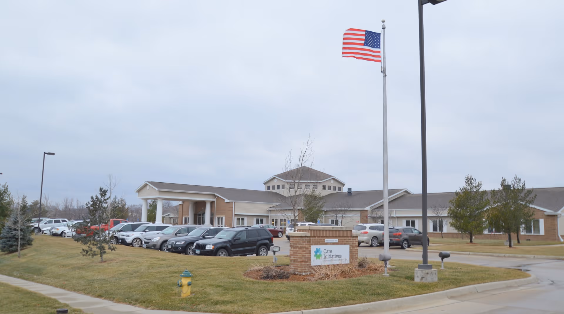 Exterior view of Northern Mahaska Specialty Care facility showing a single-story building with a covered entrance, a parking lot with several cars, a flagpole with an American flag, and a sign that reads 'Care Initiatives'. The sky is overcast and there is a grassy area with small trees and a fire hydrant in the foreground.