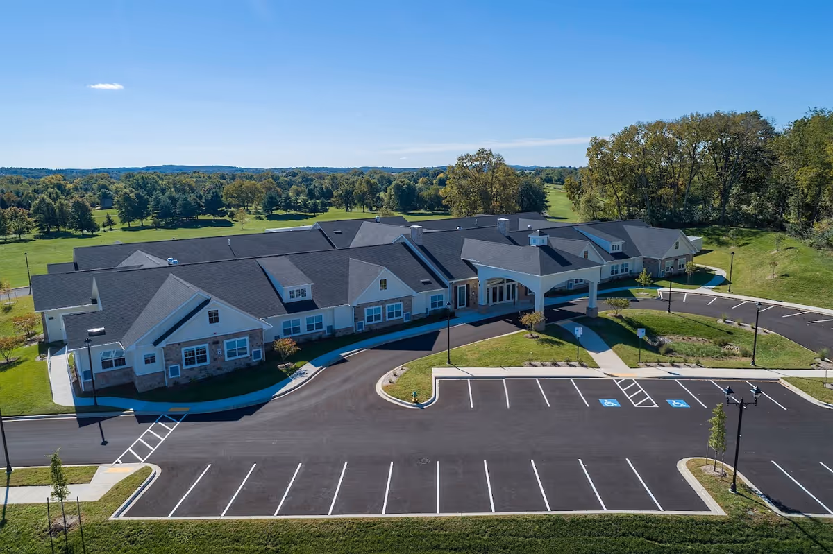 Aerial view of a single-story memory care community with a covered entrance, empty parking lot, and surrounding lawns and trees.