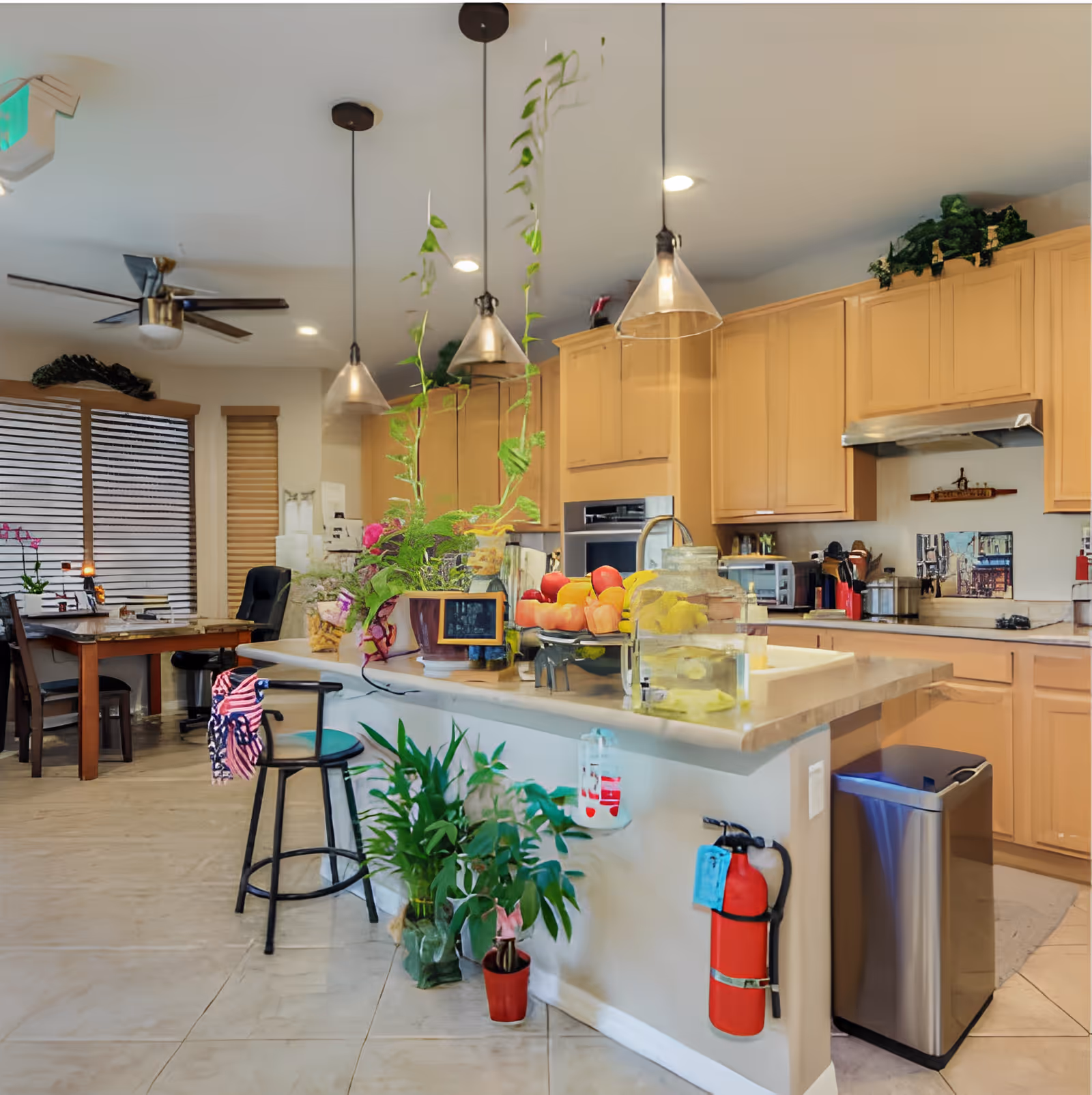 A bright kitchen area with light wood cabinets, a countertop island with hanging pendant lights, various plants, a bowl of fruit, and a fire extinguisher mounted on the side. In the background, there is a dining table with chairs and a ceiling fan.