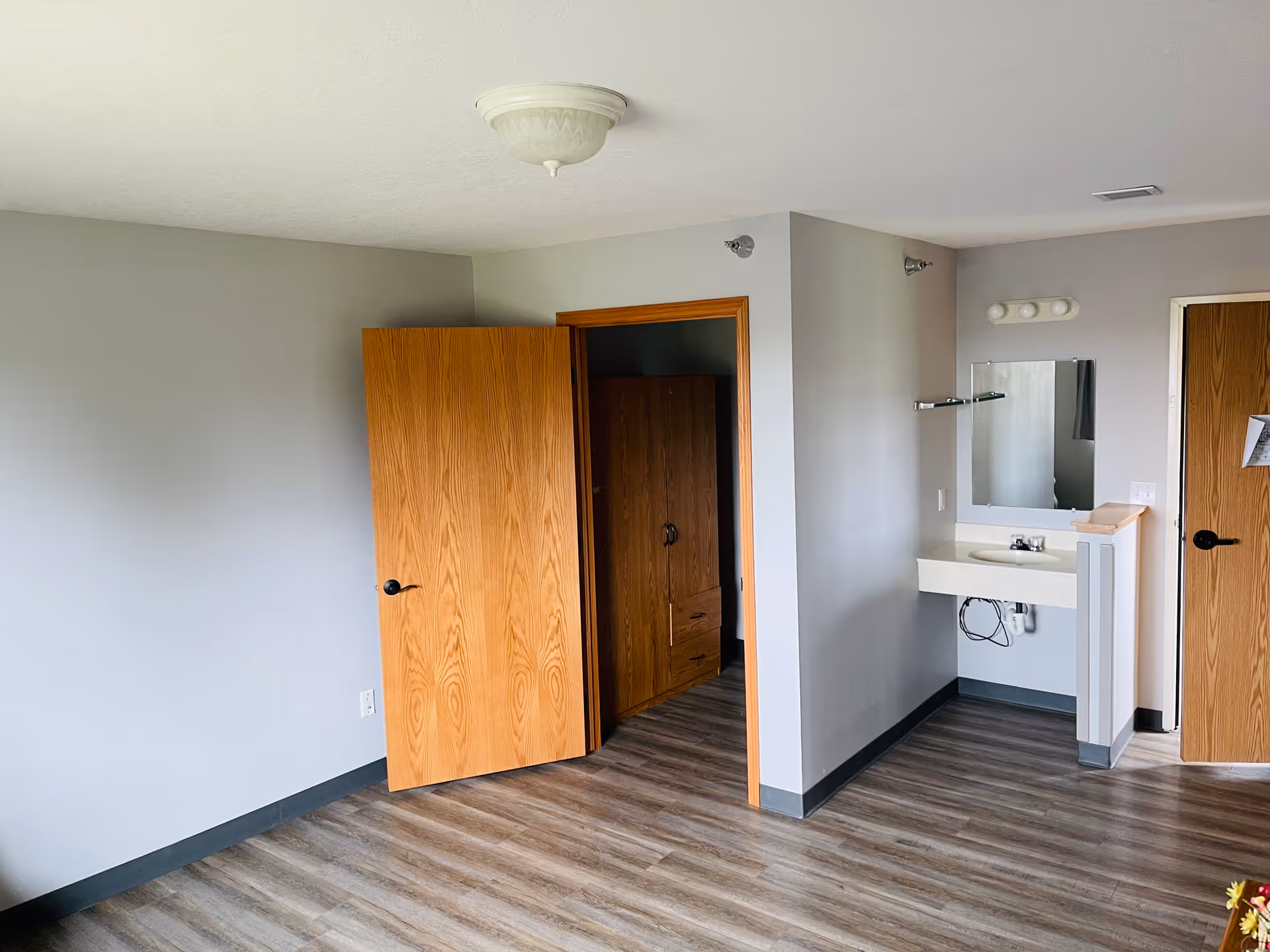 An empty room with light gray walls and wood-patterned flooring. There is a wooden door partially open revealing a closet with a wooden wardrobe inside. On the right side, there is a small sink with a mirror above it and a light fixture with three bulbs. Another closed wooden door is visible on the far right.