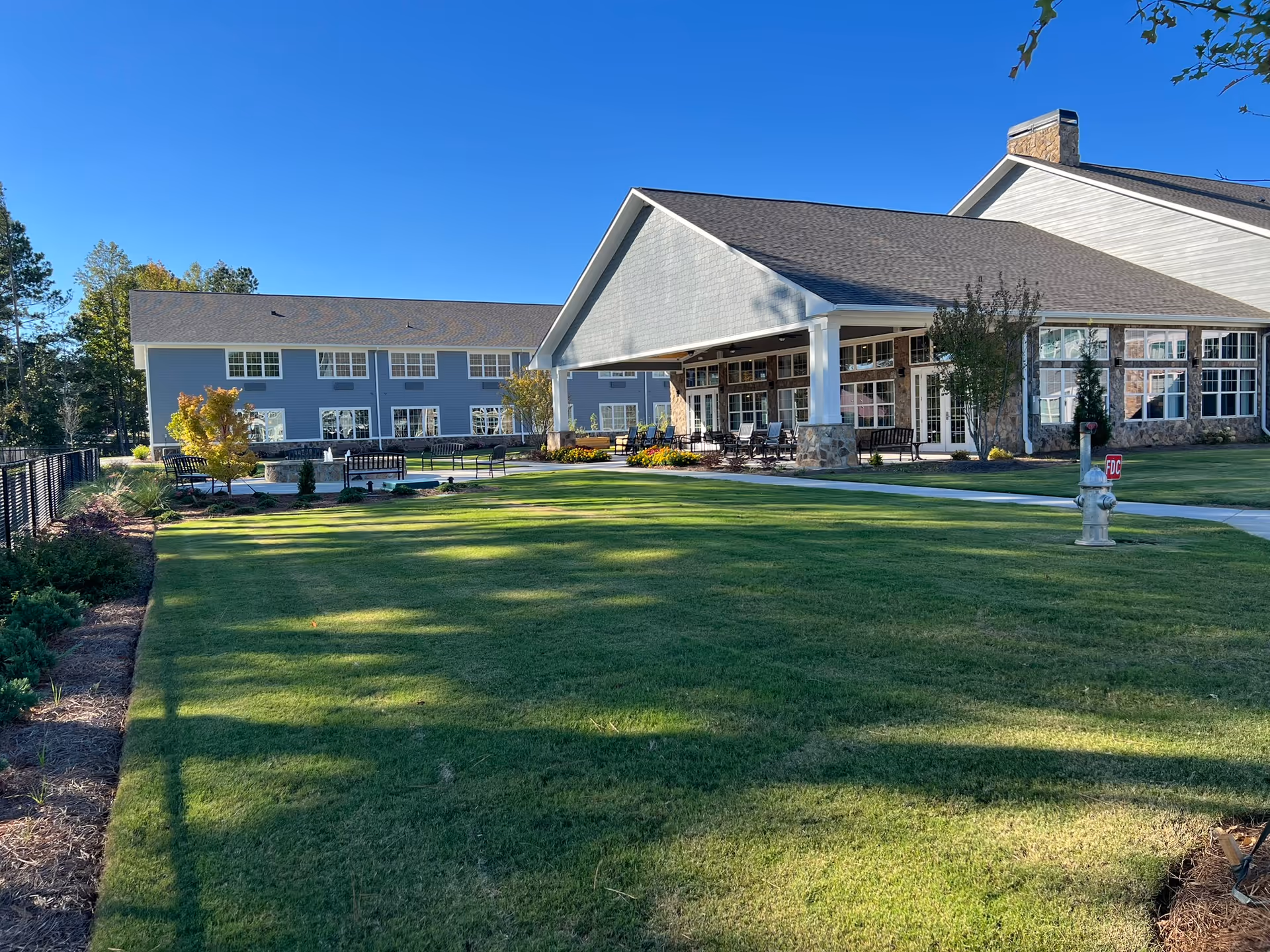 Exterior view of Manor Lake Assisted Living, Memory Care & Independent Living - Hickory Flat, showing a large building with blue siding and white trim, a covered patio area with seating, well-maintained green lawn, trees, and clear blue sky.