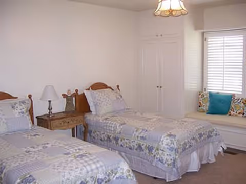 Two single beds with floral quilts, nightstand, lamp, and a window bench in a bright bedroom.