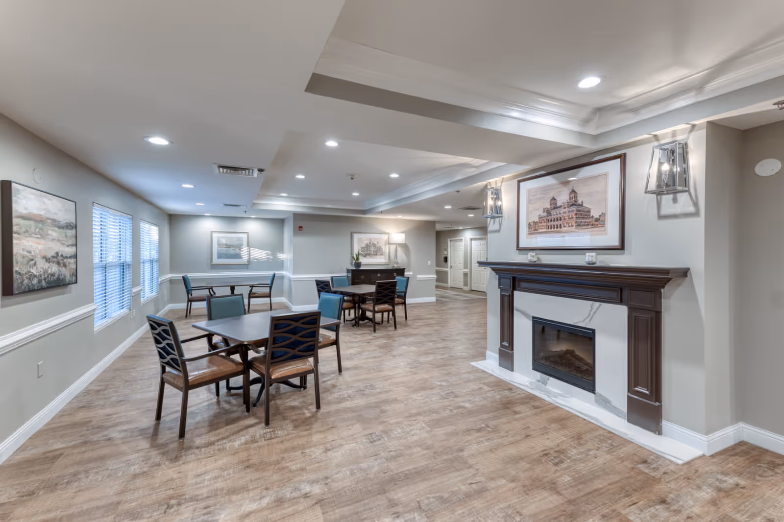 A spacious and well-lit common area with wooden flooring, several tables and chairs arranged for seating, a decorative fireplace with a framed artwork above it, and soft wall lighting. The walls are painted in a light gray color and adorned with additional framed pictures. The ceiling features recessed lighting and crown molding.