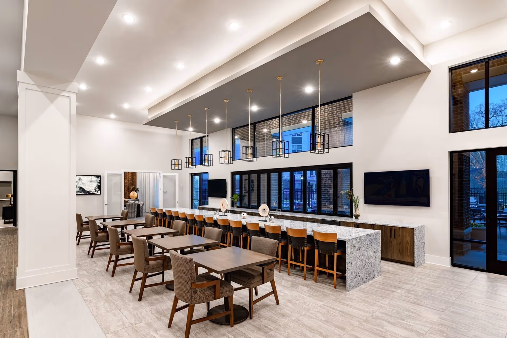 Spacious modern communal dining area featuring multiple tables and chairs, a long marble island with bar stools, pendant lighting, and large windows.