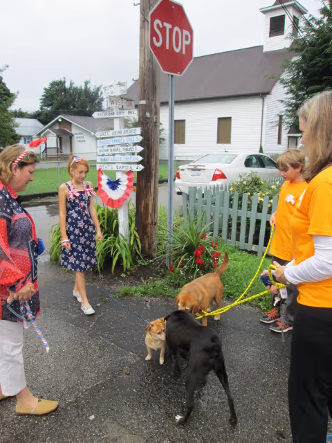 A group of people, including two women and two children, standing on a street corner with three dogs on leashes. There is a stop sign and a wooden post with multiple directional signs. A white church building and a green picket fence are visible in the background.
