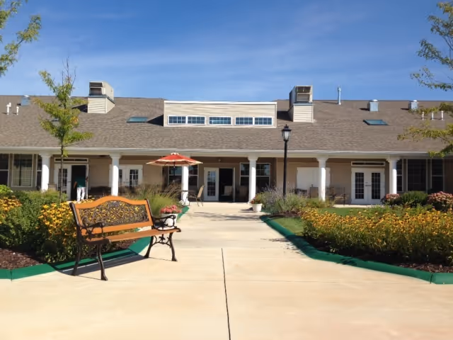 Front entrance of a single-story senior living building with a bench, landscaped walkway, and covered porch under a clear sky.