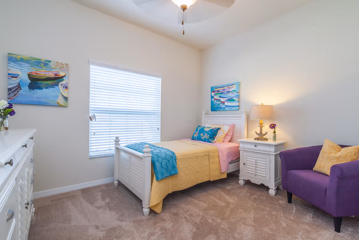 Bright furnished bedroom with a twin bed, nightstand and lamp, dresser, purple armchair, and boat-themed artwork.