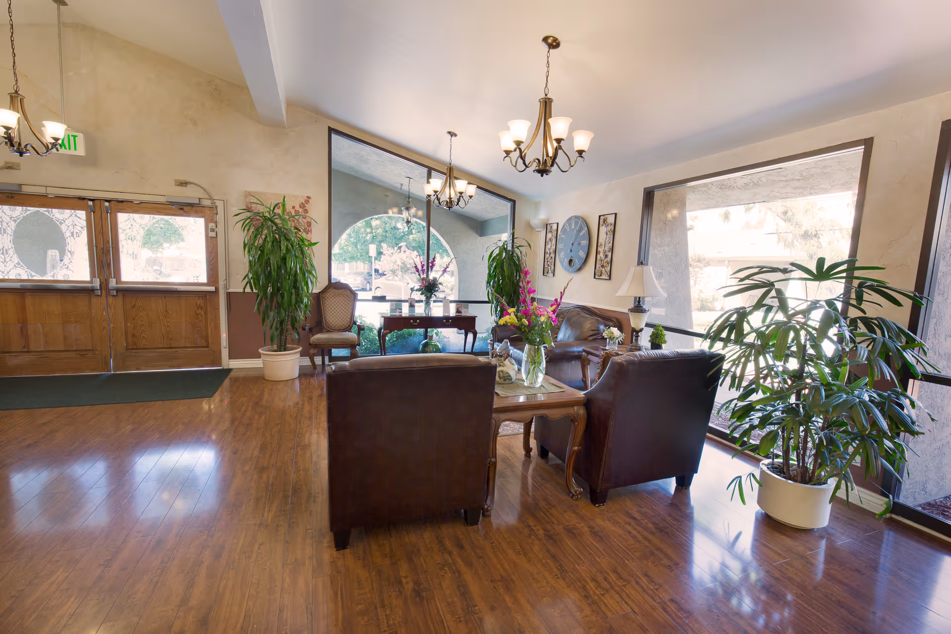A bright and welcoming living room area in Windsor Hampton Care Center featuring wooden floors, leather armchairs, a coffee table with a vase of flowers, large windows letting in natural light, potted plants, a wall clock, and decorative wall art.