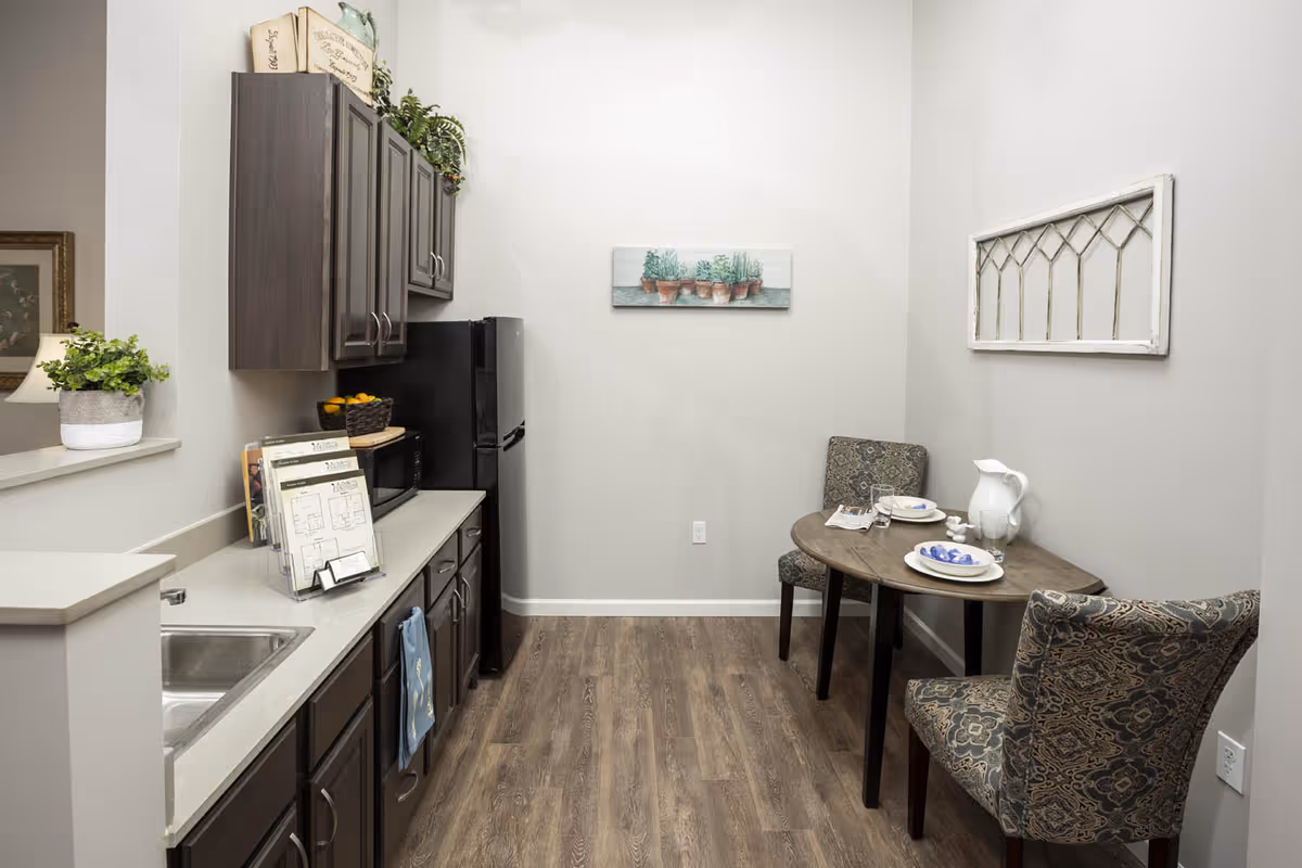 Small kitchenette with dark cabinets, sink and refrigerator on the left and a round dining table with two patterned chairs on the right.