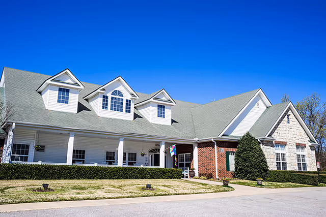 Exterior view of a single-story senior living facility building with a combination of white siding, red brick, and stone facade under a clear blue sky. The building features multiple windows, a covered porch with white columns, and a well-maintained lawn with shrubs and small trees.