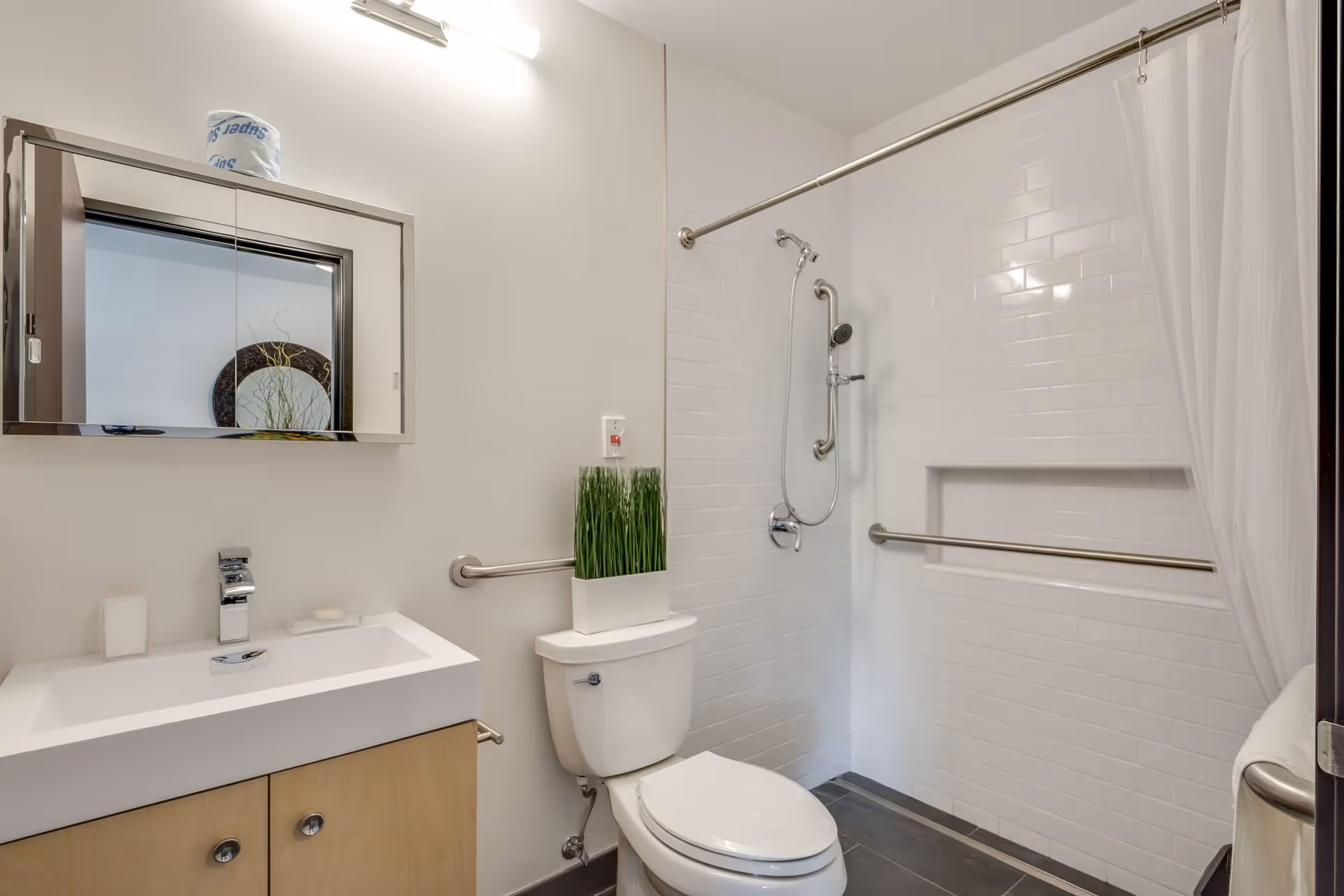 A clean and modern bathroom featuring a white toilet with a rectangular planter of green grass on top, a white sink with a chrome faucet and soap dish, a mirrored medicine cabinet above the sink, and a walk-in shower with white subway tiles, a handheld showerhead, grab bars, and a white shower curtain.