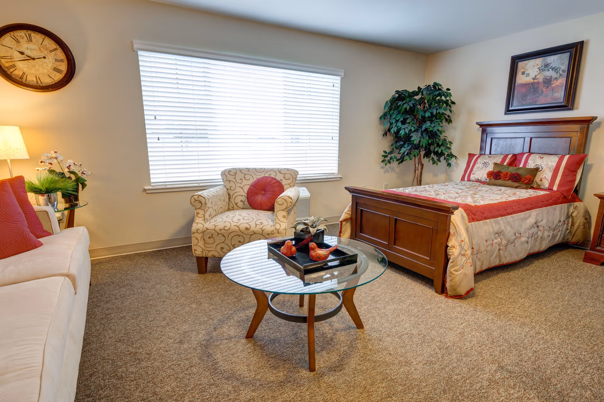 A cozy bedroom with a wooden bed featuring a red and beige floral bedspread, a matching pillow, and a framed artwork above the headboard. There is a patterned armchair with a red round pillow, a glass coffee table with decorative items, a white sofa with red pillows, a floor lamp, a large wall clock, and a potted plant near the corner. The room has beige carpet and a large window with white blinds.