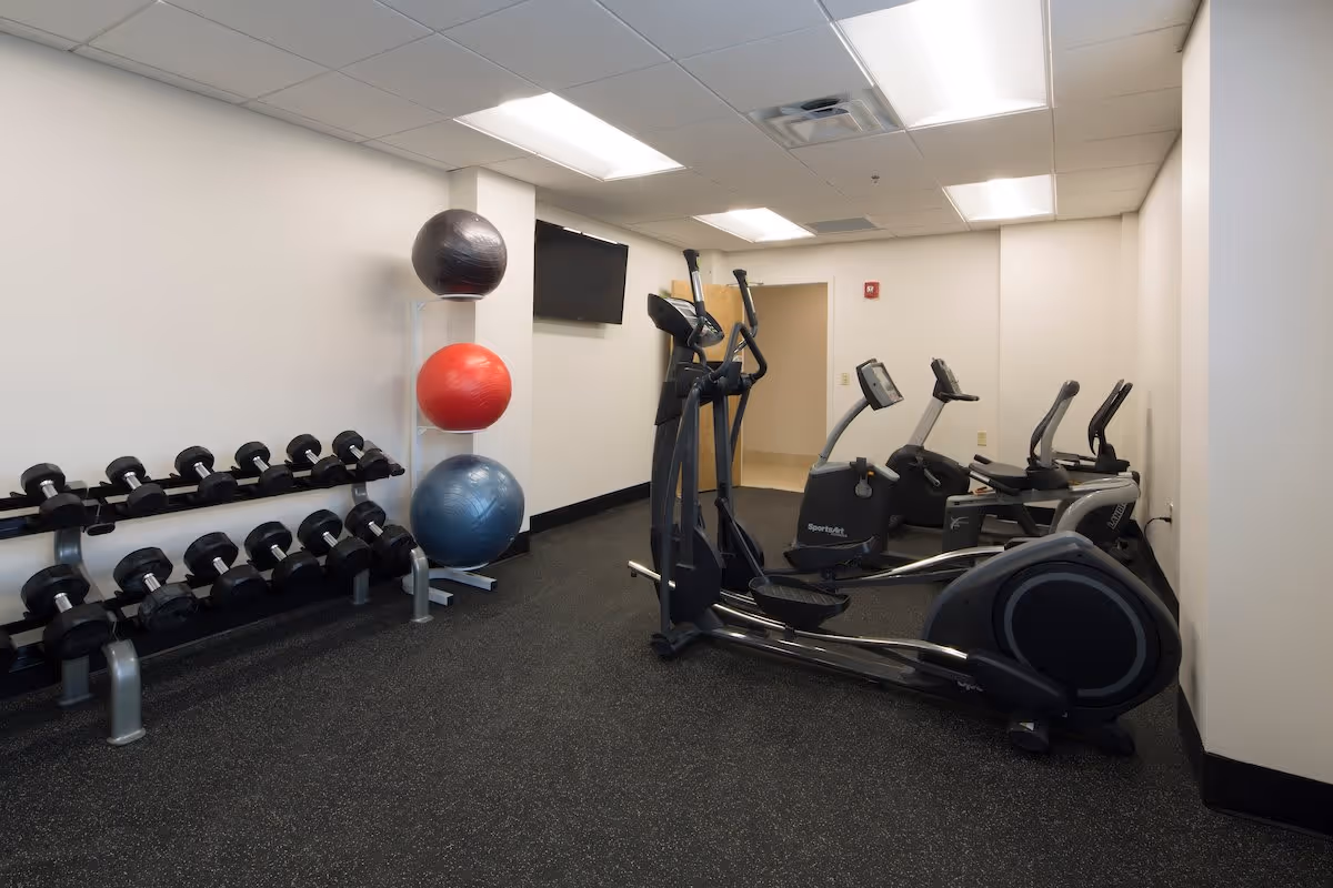 A small fitness room with exercise equipment including an elliptical machine, stationary bikes, a rack of dumbbells, and three large exercise balls stacked vertically on a stand. The room has white walls, a black speckled floor, and fluorescent ceiling lights. A flat-screen TV is mounted on the wall.