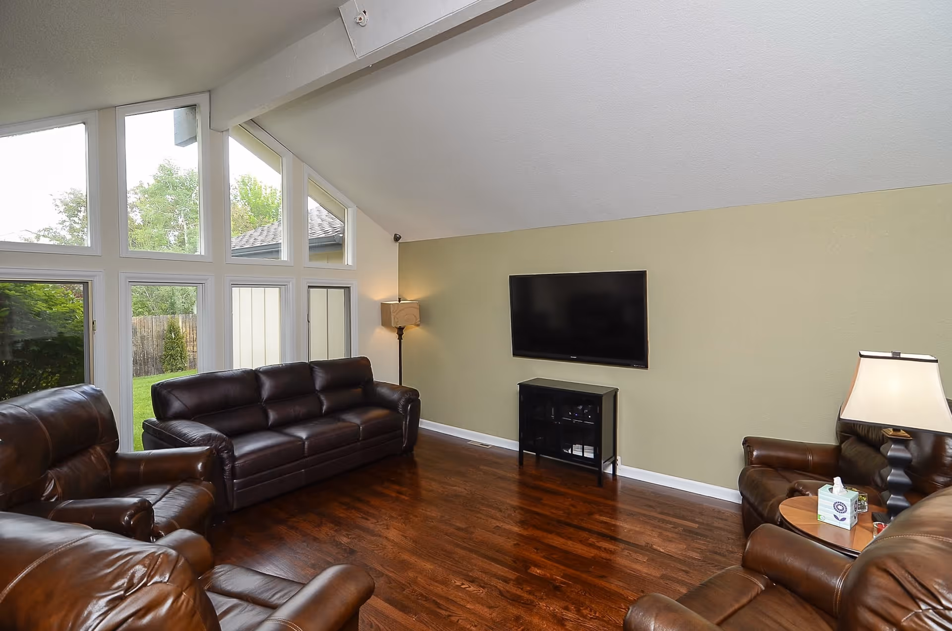 A living room with dark brown leather sofas and armchairs arranged around a wall-mounted flat screen TV. The room has large windows letting in natural light and wooden flooring. There is a floor lamp in the corner and a small side table with a lamp and tissue box.