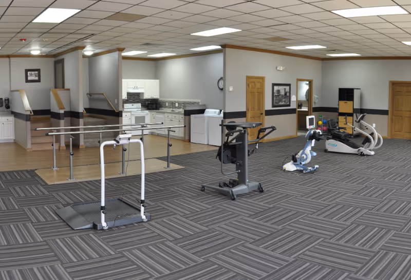 A spacious indoor rehabilitation or physical therapy room with exercise equipment including parallel bars, a stationary bike, and other fitness machines. The room has a patterned carpet floor, a drop ceiling with fluorescent lights, and a small kitchen area with white cabinets and appliances in the background. There are wooden doors and framed pictures on the walls.