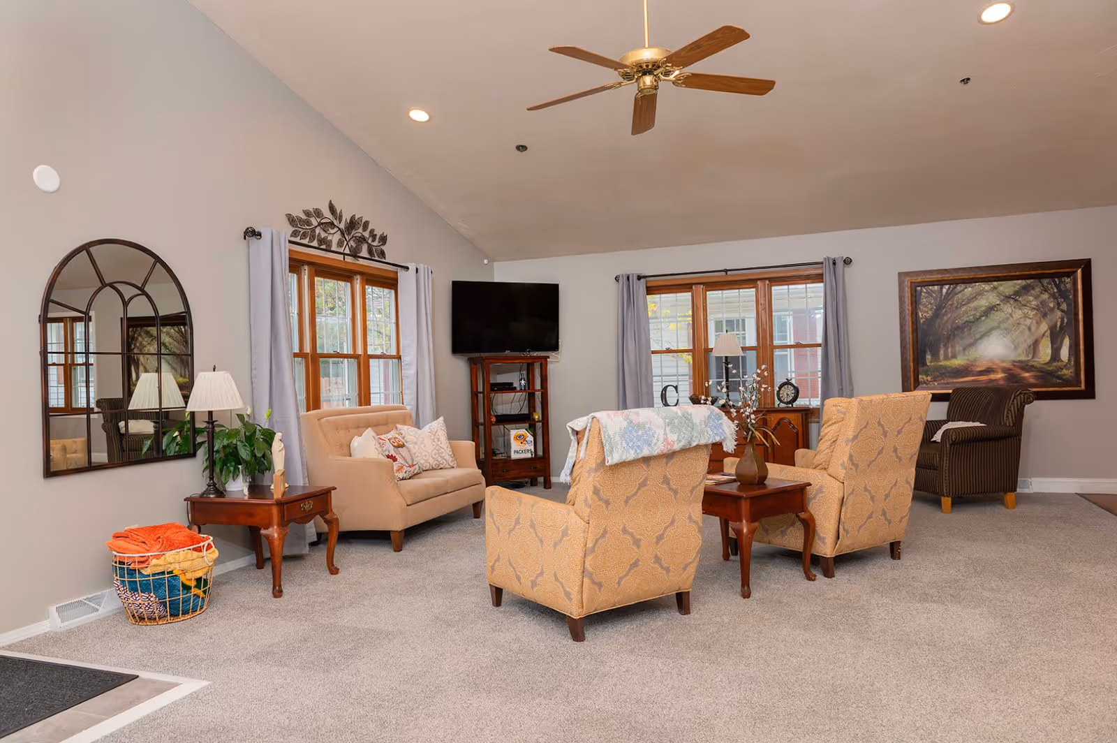 A cozy senior living room with beige carpet and light gray walls. The room features two patterned armchairs with a wooden side table between them, a loveseat with decorative pillows, a striped armchair, and a wooden TV stand with a flat-screen TV. There are two windows with light gray curtains, a large wall mirror, a table lamp, a plant, and a basket with colorful blankets. A framed painting of a tree-lined path hangs on the wall.
