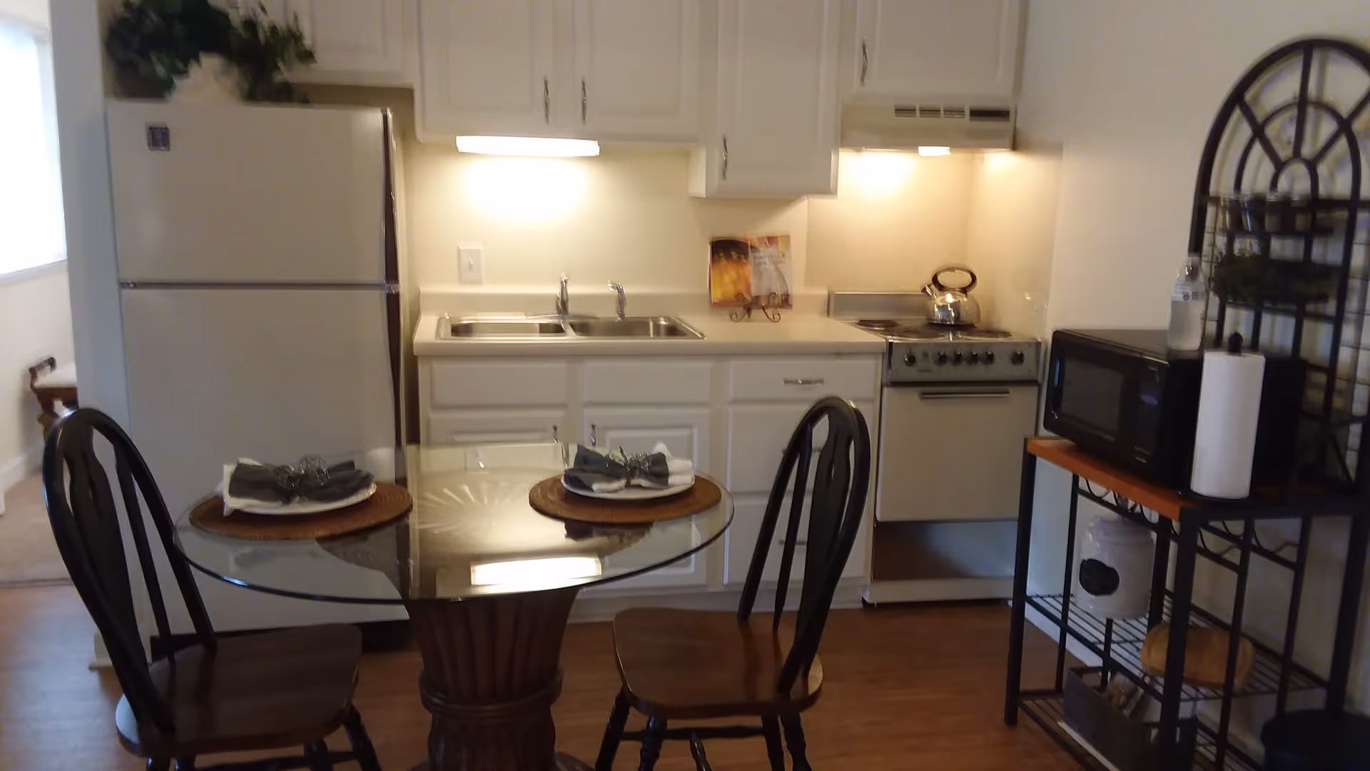 A small kitchen area with white cabinets, a double sink, a white refrigerator, and a white stove with a kettle on it. In front of the kitchen is a round glass-top table set with two place settings, each with a plate, napkin, and silverware. To the right, there is a black metal shelving unit holding a microwave, paper towels, and other kitchen items. The floor is wooden, and the space is well-lit with under-cabinet lighting.