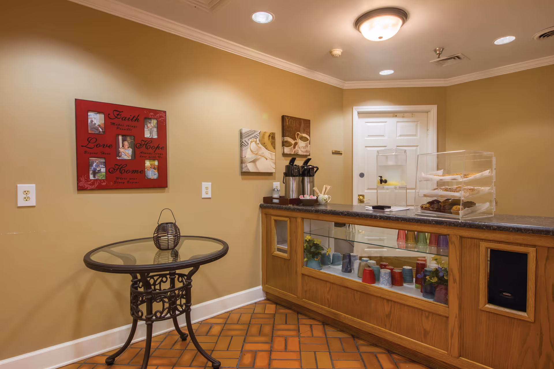 Interior view of a small refreshment area with a wooden counter displaying colorful cups and flowers inside a glass case. On the counter, there are coffee dispensers, a water dispenser with lemon slices, and a clear container with muffins and pastries. A round glass-top table with a decorative black metal base is positioned against a beige wall adorned with framed pictures and inspirational words. The floor is tiled with reddish-brown bricks, and a white door is visible in the background.