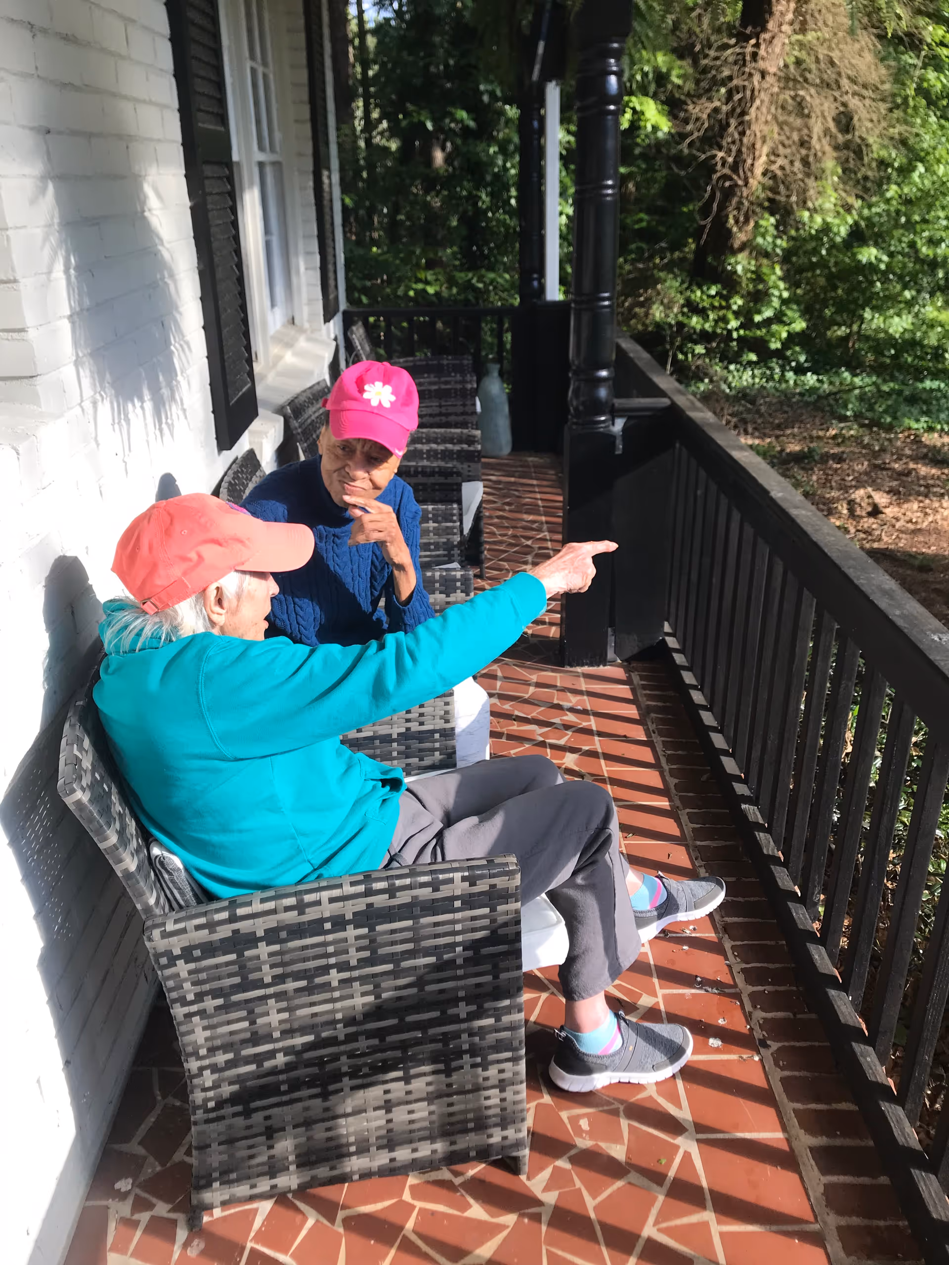 Two elderly women sitting on a porch with wicker chairs. One woman wearing a pink cap and blue sweater is listening while the other woman in a coral cap and teal jacket is pointing towards something outside the porch. The porch has a tiled floor and black railing with greenery visible beyond.