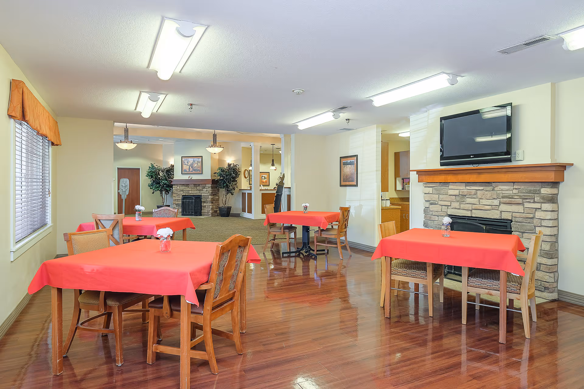 Dining/common room with tables covered in red tablecloths, wooden chairs, a stone fireplace and a wall-mounted TV.