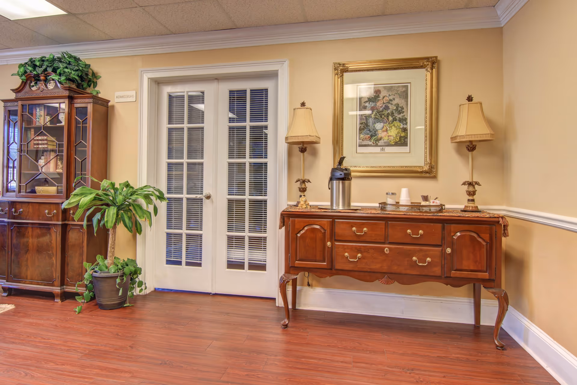 Interior view of a room with wooden flooring, beige walls, and white crown molding. There is a wooden cabinet with glass doors on the left, a potted plant in front of it, and double glass doors labeled 'Admissions' in the center. On the right, there is a wooden sideboard with two table lamps, a coffee dispenser, and a tray with cups and condiments. Above the sideboard hangs a framed floral painting.