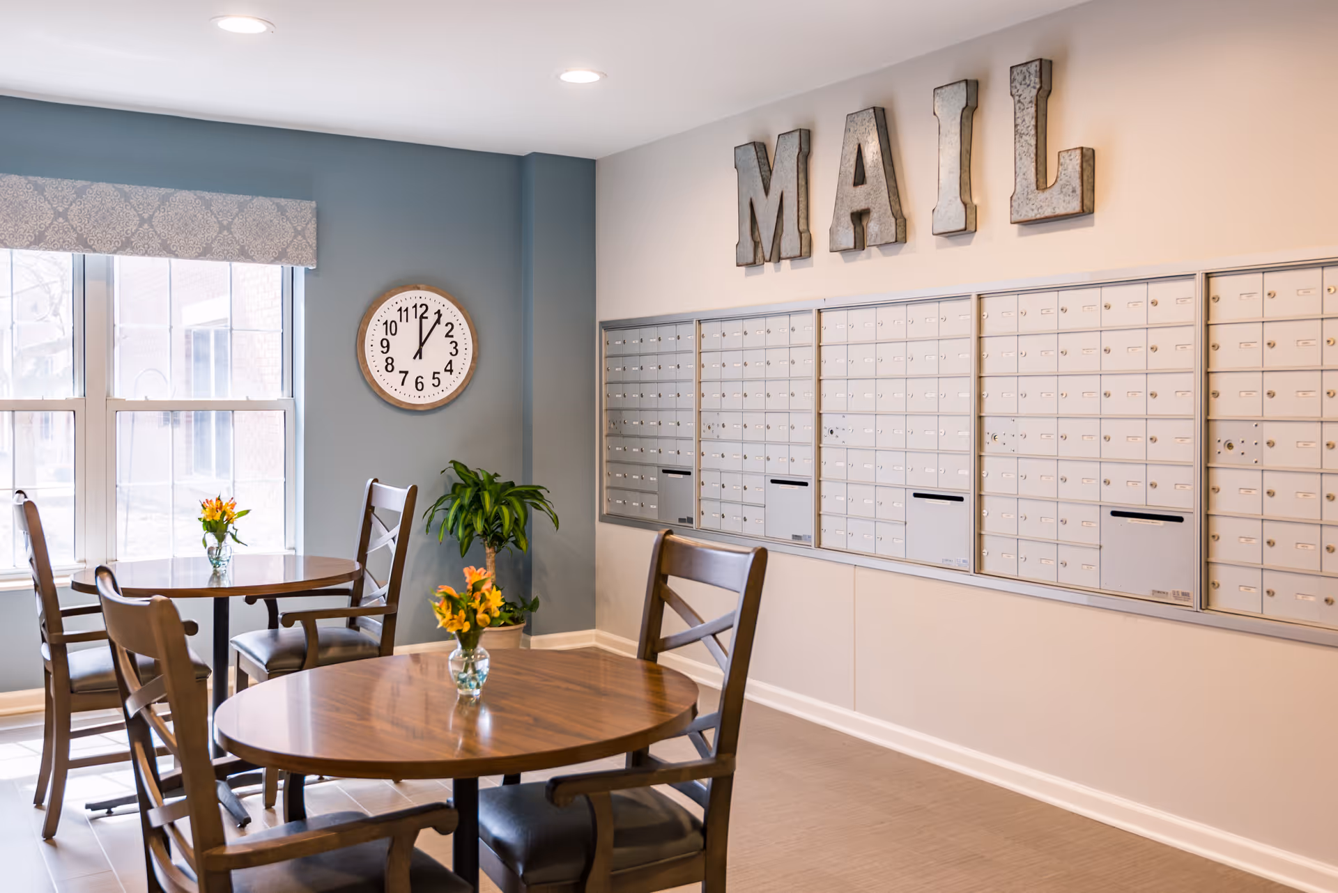 Mailroom with rows of metal mailboxes on the wall, round wooden tables and chairs, a clock, and decorative 'MAIL' letters.