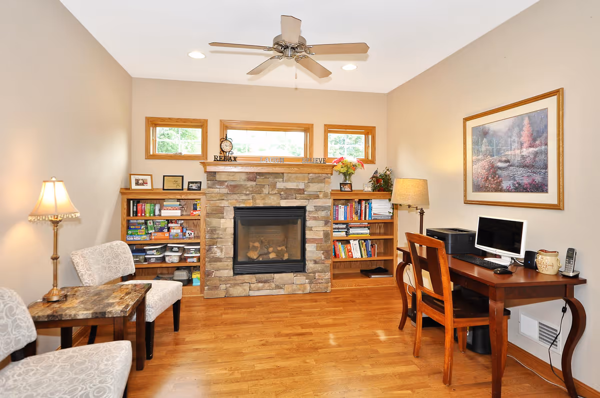A cozy room with a stone fireplace centered between two wooden bookshelves filled with books and games. There are two patterned chairs with a small table and lamp on the left side, and a wooden desk with a computer, printer, and chair on the right side. The walls are beige, and there is a ceiling fan above. A framed landscape painting hangs on the right wall.