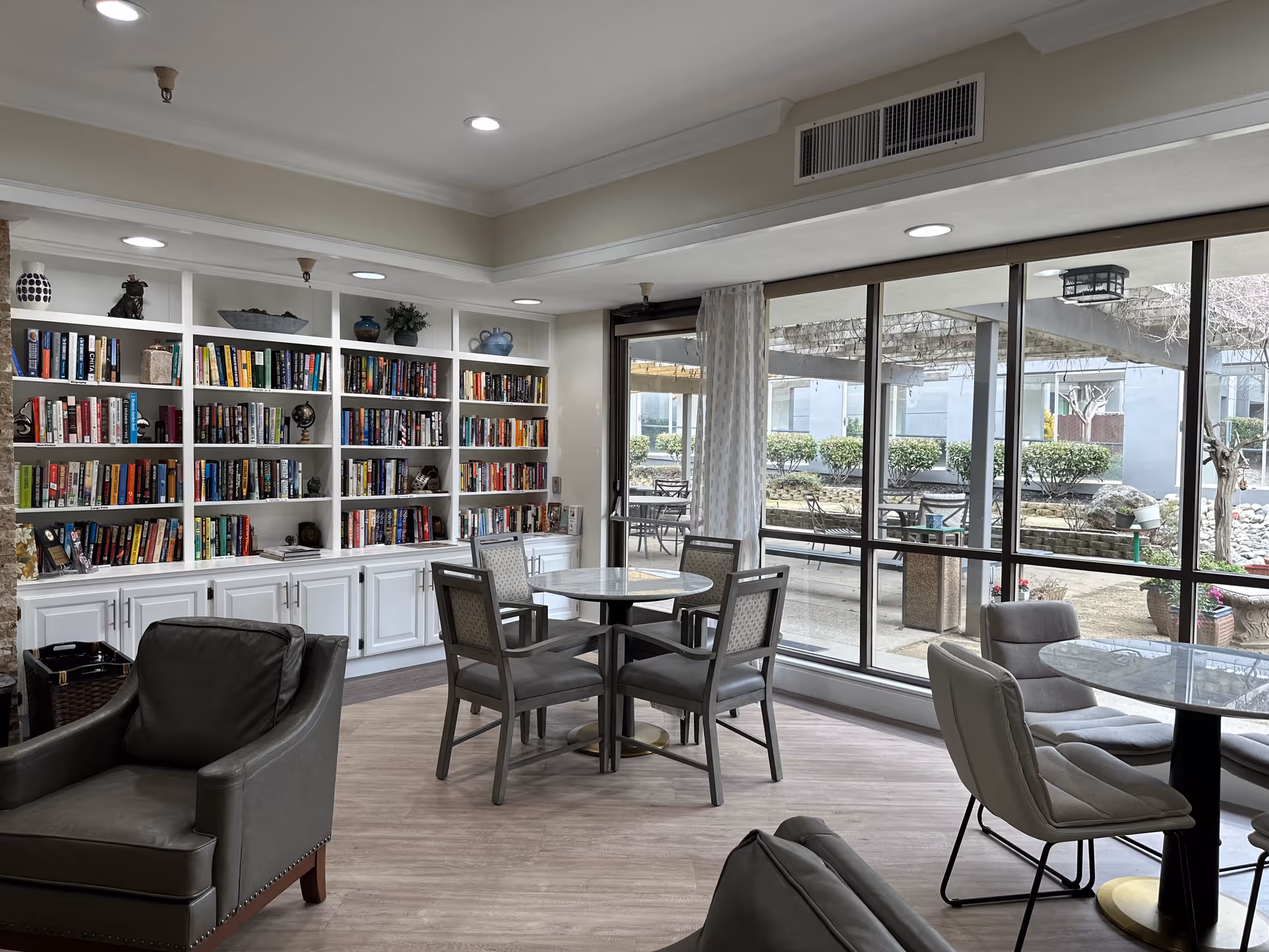 A cozy senior living common area with a large white bookshelf filled with books along one wall. There are several round tables with chairs arranged around them, and comfortable armchairs in the foreground. Large windows provide a view of an outdoor patio area with more seating and greenery.