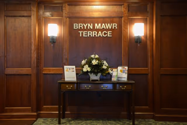 Wood-paneled interior wall with a table holding a floral arrangement and brochures beneath a sign that reads 'BRYN MAWR TERRACE' flanked by two wall sconces.