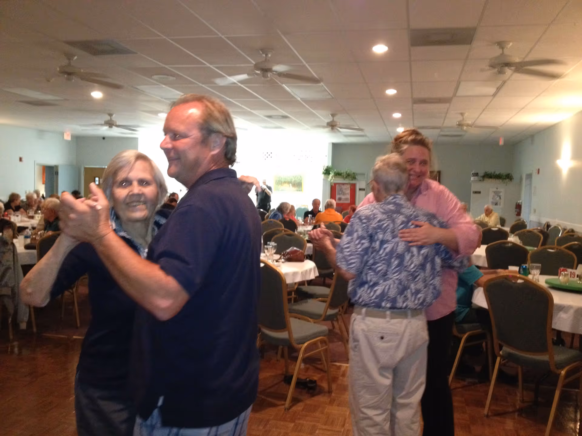 Two couples dancing in a dining room with round tables and chairs. Other people are seated at tables in the background, and the room is well-lit with ceiling lights and fans.