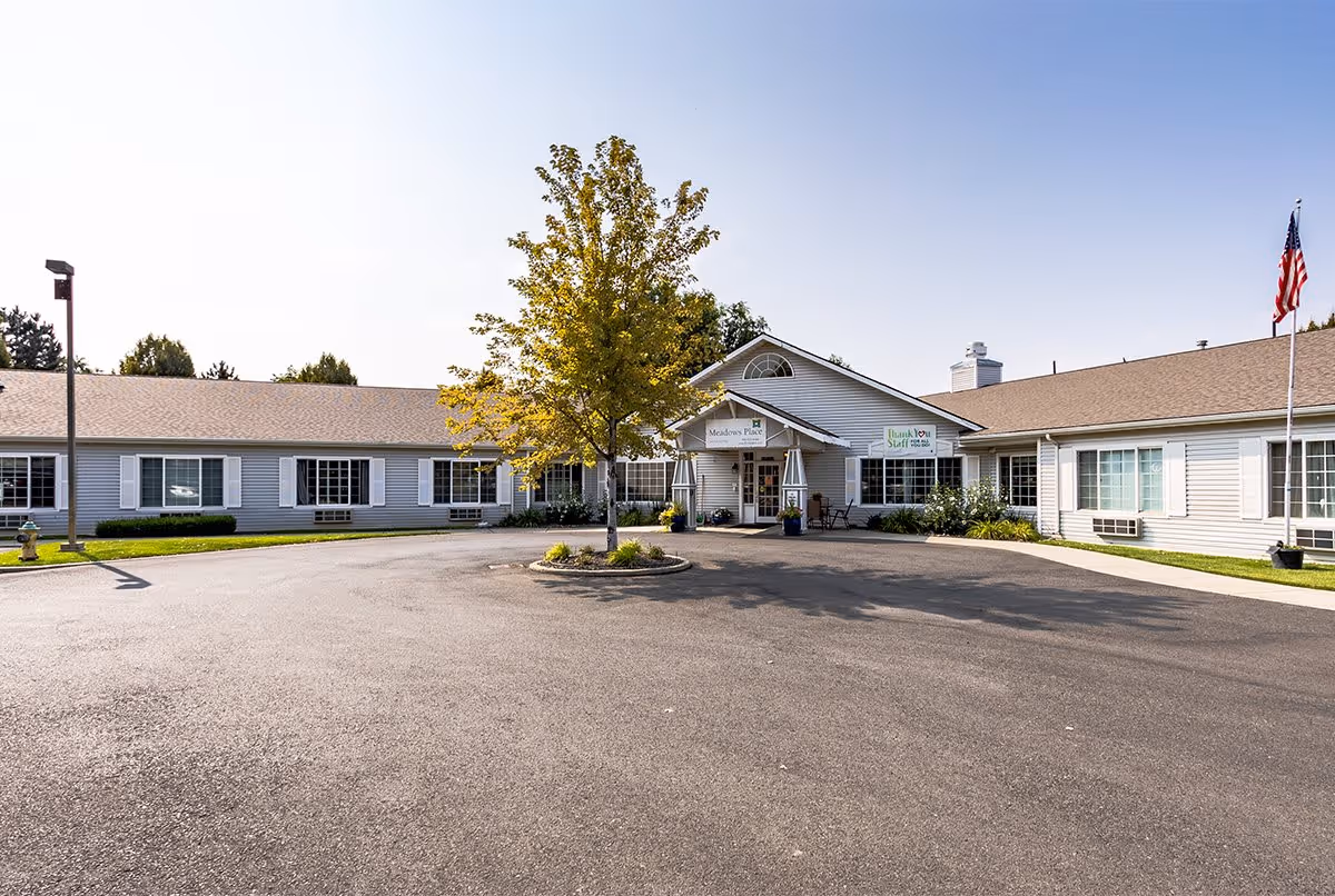 Exterior view of a single-story senior living facility building with light gray siding and white trim. There is a paved driveway in front with a small landscaped island featuring a tree. The entrance has a covered porch with a sign that reads 'Meadows Place' and another sign thanking the staff. An American flag is visible on a flagpole to the right.