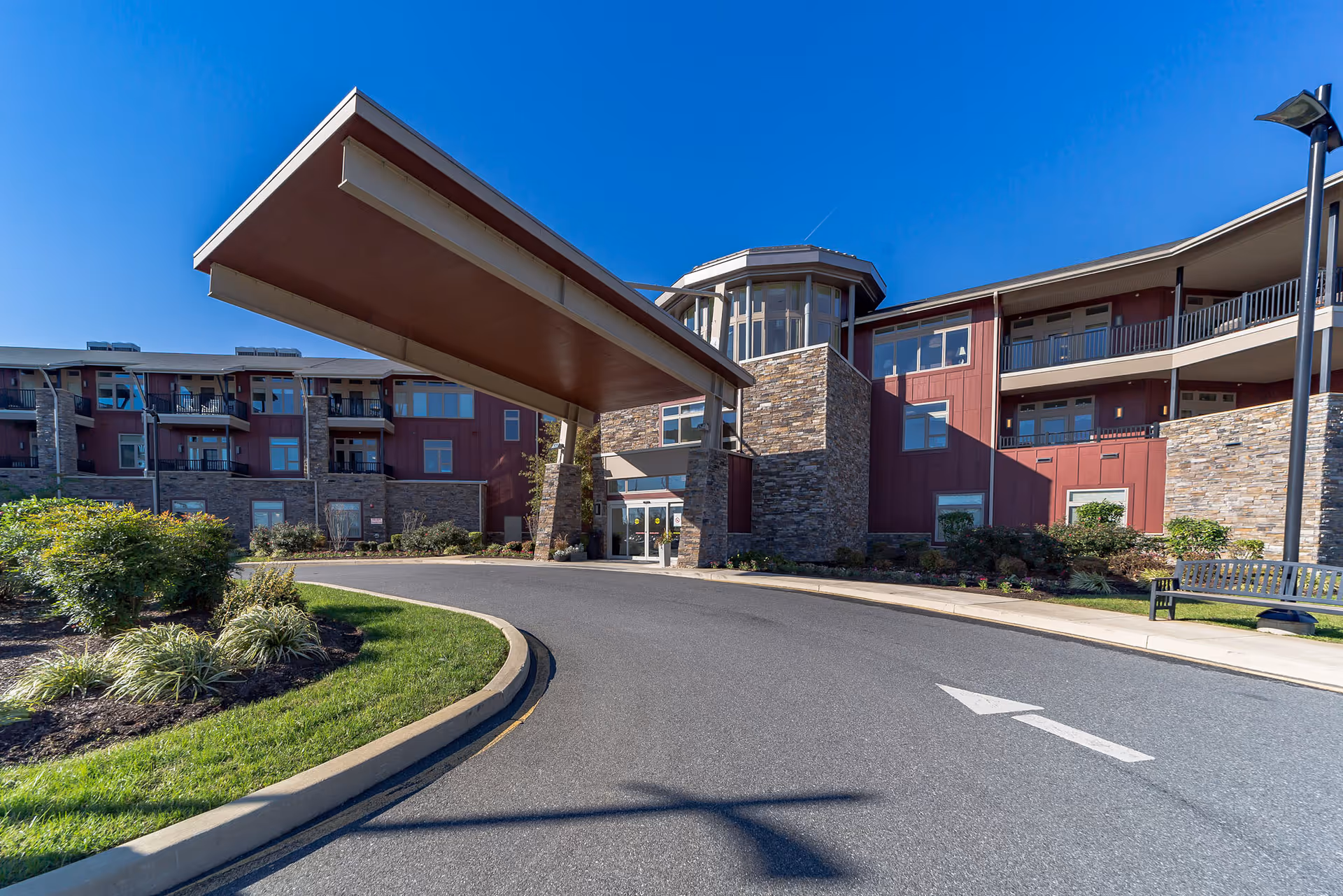 Front entrance of a senior living building with a large covered porte-cochere, circular driveway, and landscaped grounds under a clear blue sky.