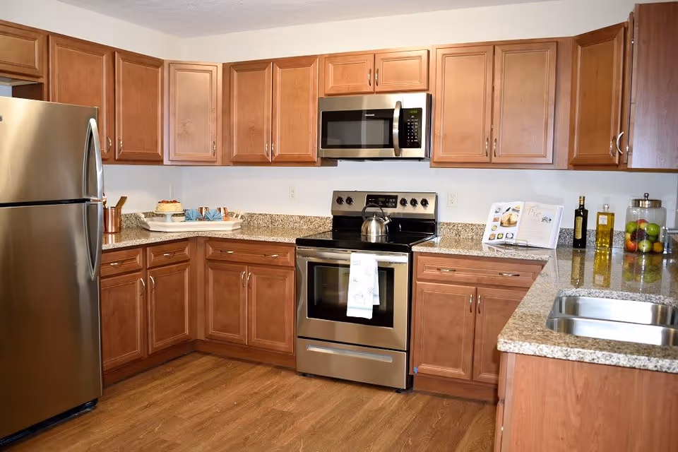 A modern kitchen with wooden cabinets, granite countertops, a stainless steel refrigerator, stove with oven, microwave, and a double sink. There are kitchen items including a kettle on the stove, a cake on a tray, a cookbook, bottles of oil, and a jar of fruit on the countertops.