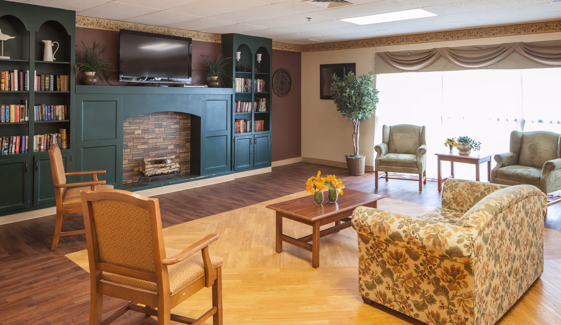A cozy living room with a floral patterned sofa, two wooden armchairs with orange cushions, and two green upholstered armchairs near a large window with beige curtains. A wooden coffee table with yellow flowers in vases is centered on a two-tone wood floor. The room features a green built-in bookshelf and fireplace with a mounted flat-screen TV above it, and potted plants on either side of the fireplace.