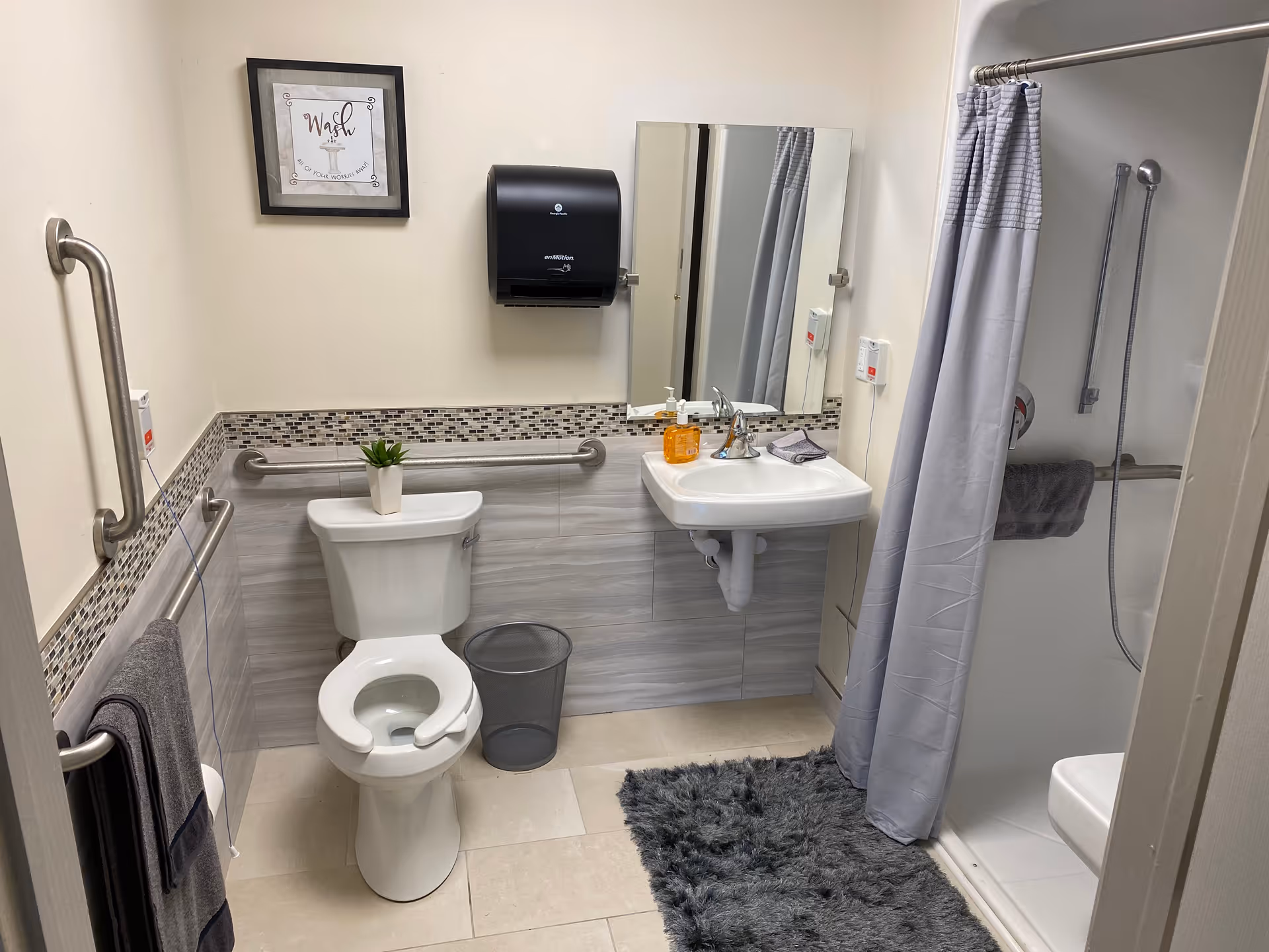 Accessible bathroom featuring a toilet, wall-mounted sink, shower with curtain, multiple grab bars, and a gray shag rug.