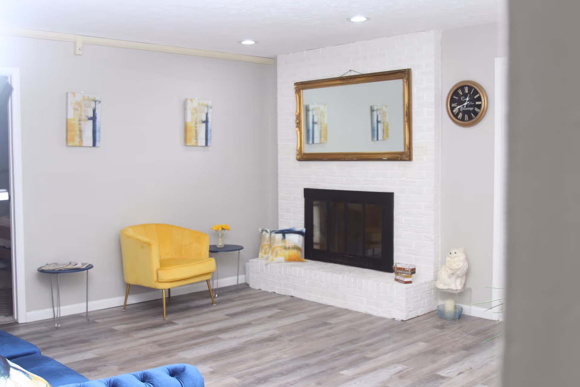 Bright living room with a yellow armchair, white brick fireplace topped by a mirror and a wall clock.