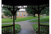 View from inside a wooden gazebo looking out onto a paved walkway and a large grassy area with trees and a multi-story brick building in the background.