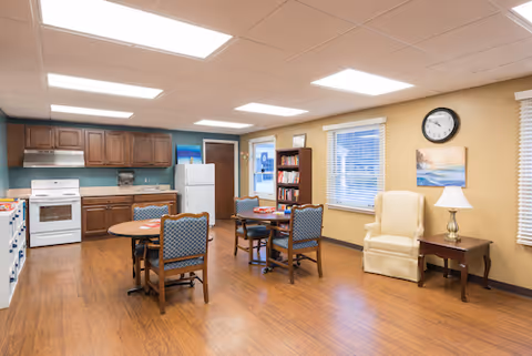 A bright room with a kitchen area featuring wooden cabinets, a white stove, and refrigerator. There are two round tables each surrounded by four blue cushioned chairs. A beige armchair sits next to a wooden side table with a lamp. The walls are light yellow with two windows, a clock, and a painting. The floor is wooden and the ceiling has recessed lighting.
