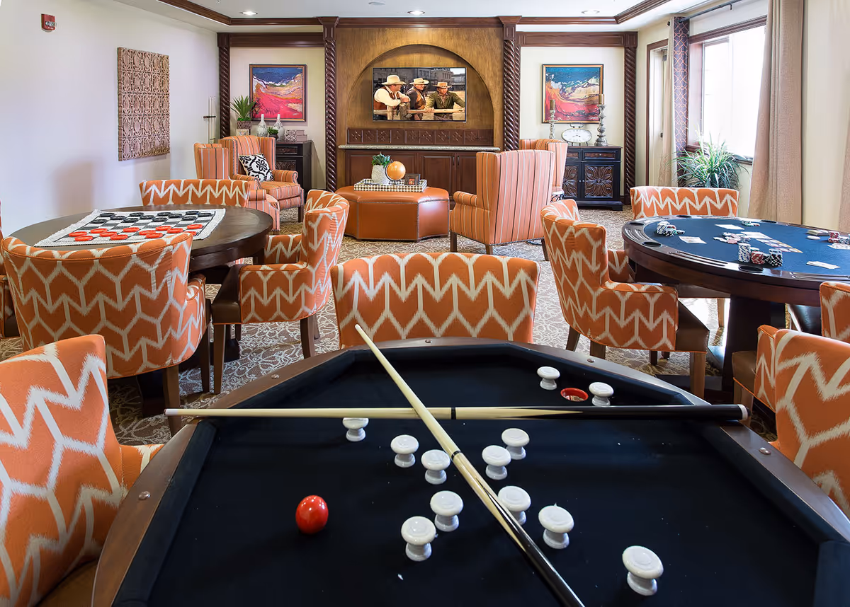 A cozy game room with a pool table in the foreground, two pool cues crossed on the table, and white game pieces scattered on the black felt. Behind the pool table are two round tables with orange patterned chairs, one table has a checkers board and the other has poker chips and cards. The room has a carpeted floor, large windows with curtains, and a wooden entertainment center with a TV showing a western scene. The walls are decorated with colorful paintings and plants are placed around the room.