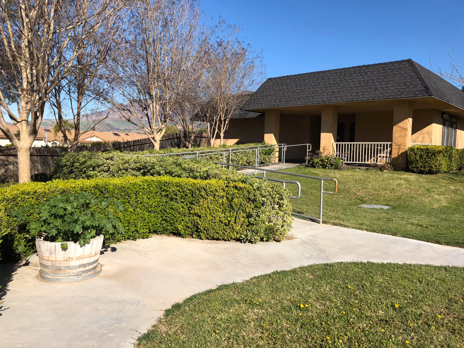 Front exterior of a single-story building with a wheelchair ramp, hedges, trees, and a paved walkway under a clear blue sky.