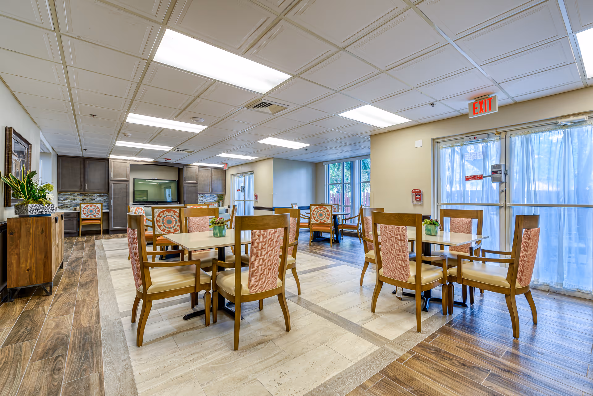 A bright and spacious dining area in a senior living facility with several wooden tables and chairs arranged neatly. The chairs have cushioned seats and backs with a patterned fabric. Large windows and glass doors with sheer curtains allow natural light to fill the room. The floor features a combination of wood and tile, and there are plants and framed artwork adding a decorative touch.