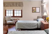 A bright and clean nursing home bedroom with two single beds covered in light green quilts. There is a wooden nightstand with a lamp and a vase of sunflowers between the beds. A cushioned armchair and a small wooden table are positioned near a window with a beige valance. A bulletin board is mounted on the wall above the nightstand.