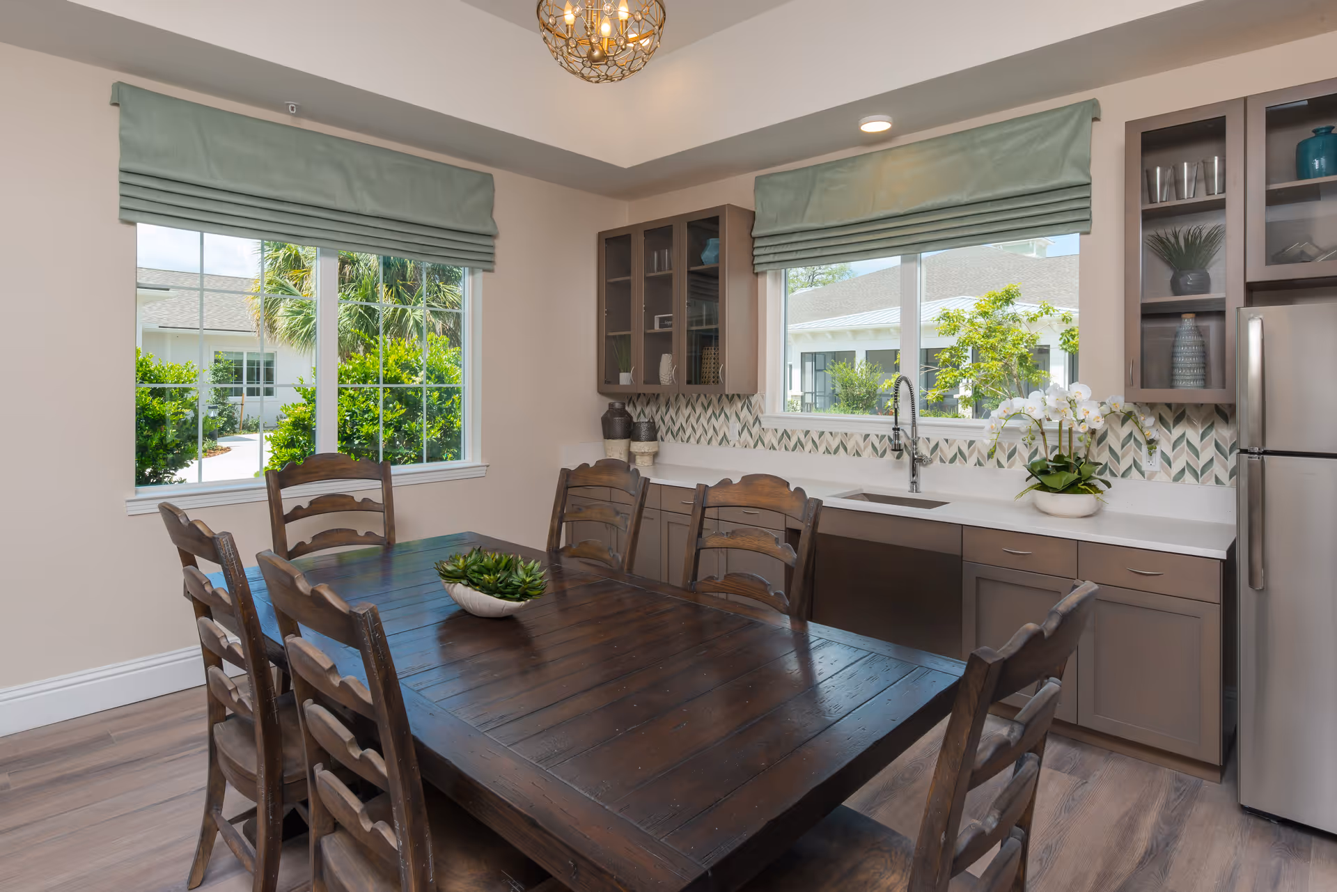 A bright kitchen and dining area featuring a dark wooden dining table with six matching chairs. The kitchen has light brown cabinets, a white countertop, a stainless steel refrigerator, and a sink under a large window with green roman shades. Decorative plants and vases are placed on the countertop and shelves. Outside the windows, greenery and parts of the building are visible.